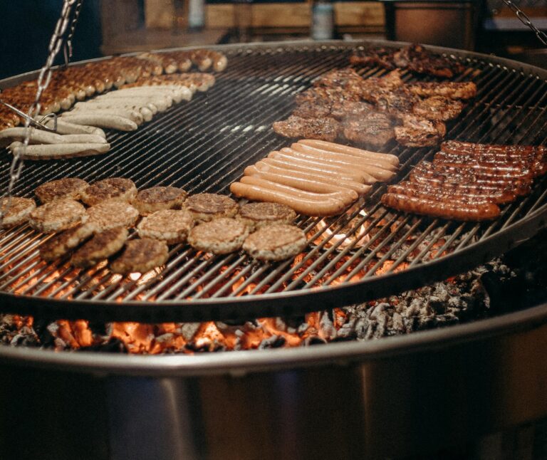 Assorted sausages and meat patties grilling over an open flame, perfect for street food at outdoor events.